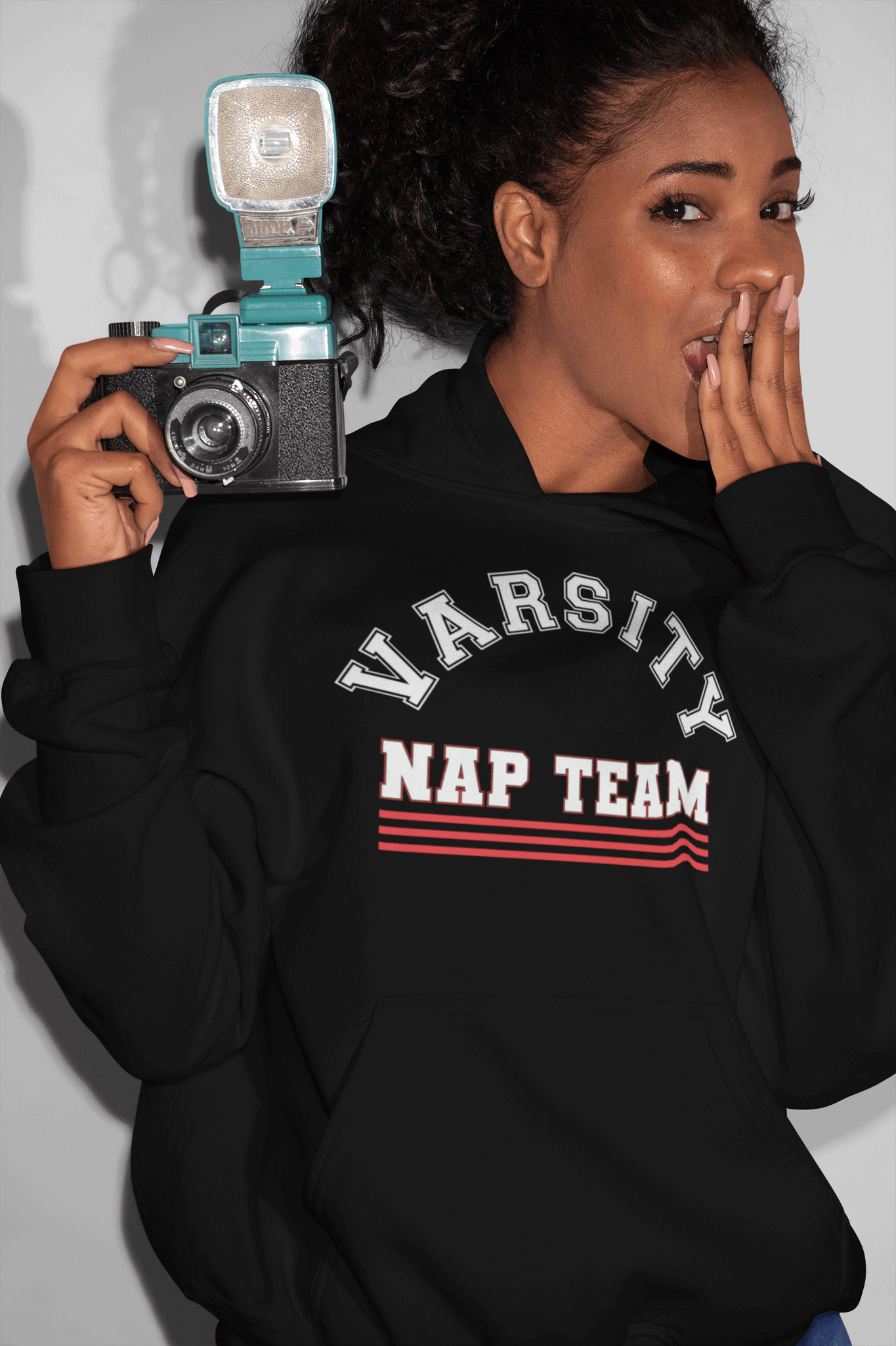 African American woman holding camera with hand over mouth wearing a black hoodie sweatshirt with Varsity Nap Team design on the front, available from the Xpert Apparel Store.