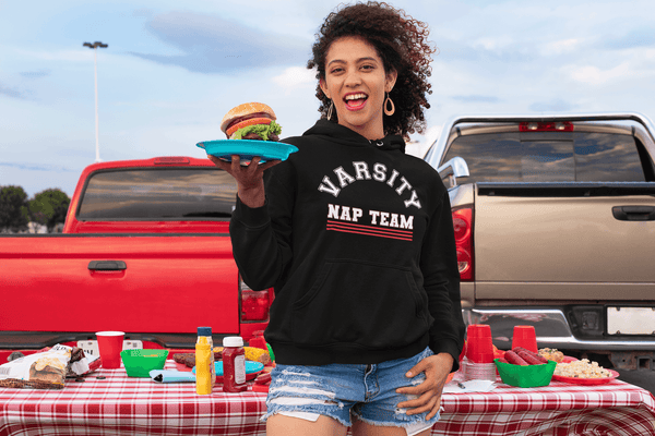Woman at tailgate party holding plate with cheese burger wearing short blue jeans and black hoodie sweatshirt with Varsity Nap Team design on the front , available from the Xpert Apparel Store.