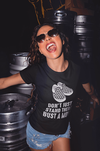 Lady having fun in a brewery, kegs of beer behind her, wearing a Don't just stand there bust a move Black T-shirt