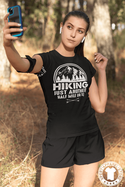 Young Lady hiking in the woods stoped to take a selfie wearing a Black T-shirt with "Hiking, just another half mile or so" design on the front. available from the Xpert Apparel Store.