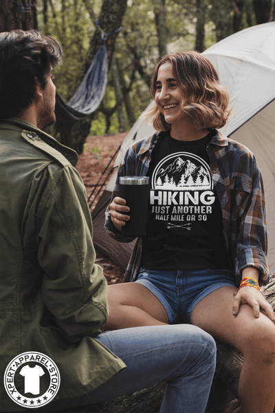 Woman Sitting on log outside her tent with a  friend wearing a flannel shirt and black t-shirt with "Hiking, just another half mile or so" design on the front. available from the Xpert Apparel Store.